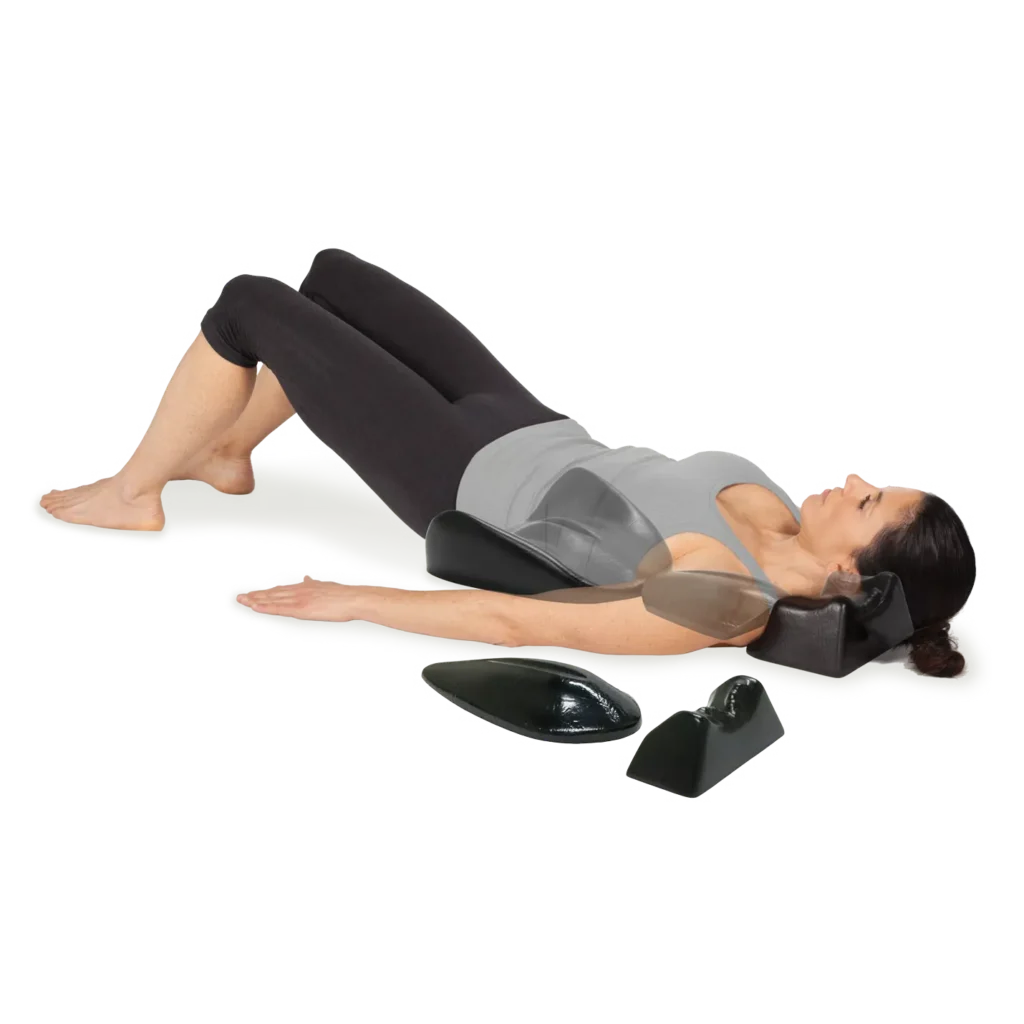 Woman lying on her back with knees bent and feet flat, performing a bridge-like exercise on a mat with a foam neck support and small leg/foot wedges nearby.